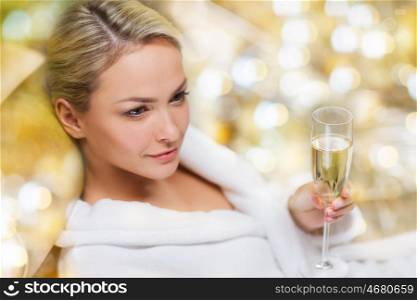 people, and relaxation concept - beautiful young woman in white bath robe lying on chaise-longue and drinking champagne at spa over holidays lights background