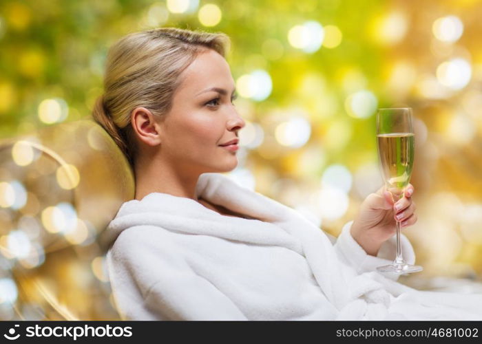 people and relaxation concept - beautiful young woman in white bath robe lying on chaise-longue and drinking champagne at spa over holidays lights background. beautiful young woman drinking champagne at spa