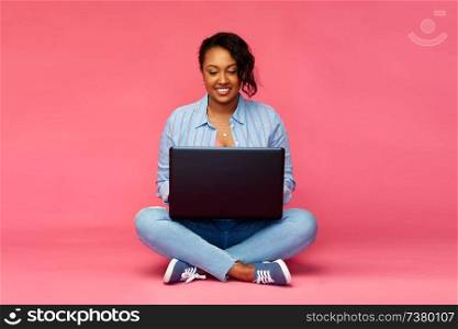 people and technology concept - happy african american young woman with laptop computer sitting on floor over pink background. happy african american woman with laptop computer
