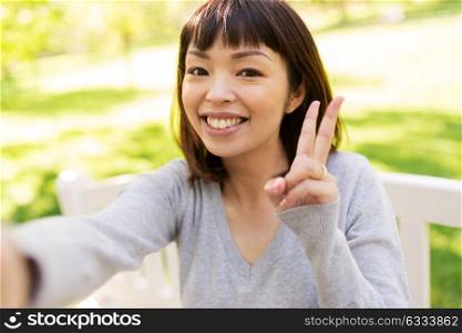 people and technology concept - happy smiling asian woman taking selfie at park and showing peace hand sign. happy asian woman taking selfie and showing peace