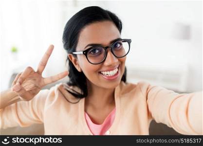 people and technology concept - happy smiling indian woman taking selfie at home and showing peace hand sign. smiling woman taking selfie and showing v sign