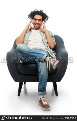 people and technology concept - happy smiling young man in headphones and glasses sitting in chair and listening to music over white background. man in headphones listening to music in chair