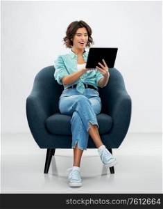 people and technology concept - portrait of smiling young woman in turquoise shirt and jeans with tablet pc computer sitting in modern armchair over grey background. happy woman with tablet pc sitting in armchair