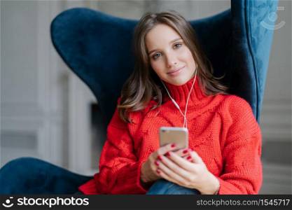 People and technology concept. Satisfied female meloman enjoys favourite music, listens audio record, dressed in red sweater, surfes in playlist, sits in armchair in living room, has spare time