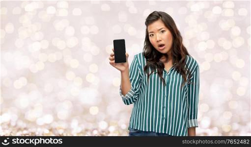 people and technology concept - shocked asian young woman with smartphone over festive lights on beige background. shocked asian woman with smartphone over lights
