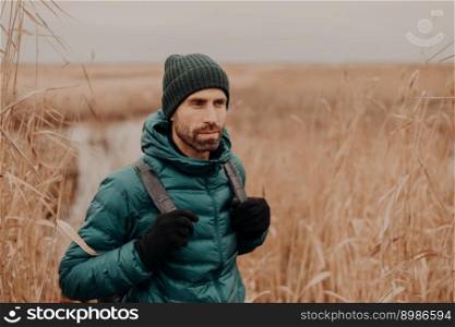People and tourism concept. Pensive unshaven handsome young man with rucksack, dressed in stylish clothes, focused into distance, poses against outdoor field background. Lifestyle. Adventure