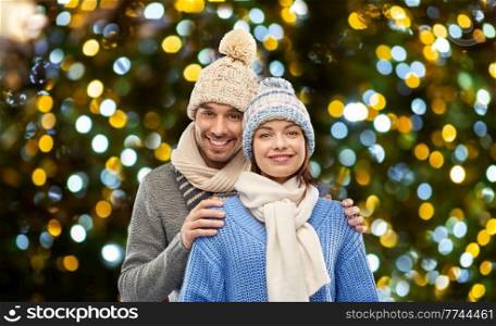 people and winter holidays concept - happy couple in knitted hats and scarves over christmas lights background. happy couple in winter clothes christmas lights