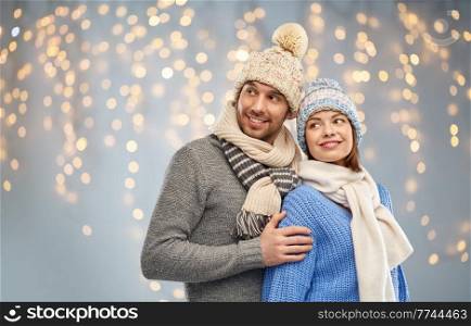 people and winter holidays concept - happy couple in knitted hats and scarves over christmas lights background. happy couple in winter clothes