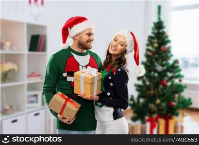 people and winter holidays concept - happy couple in santa hats with gifts in ugly sweaters over christmas tree at home background. happy couple in ugly sweaters with christmas gifts