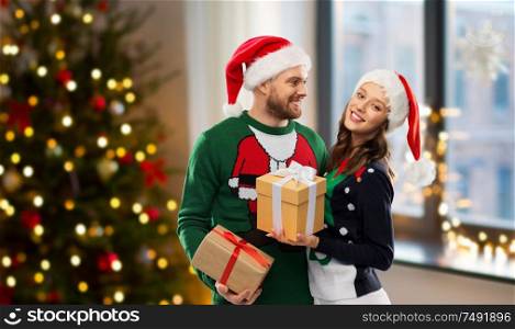 people and winter holidays concept - happy couple in santa hats with gifts in ugly sweaters over christmas tree at home background. happy couple in ugly sweaters with christmas gifts