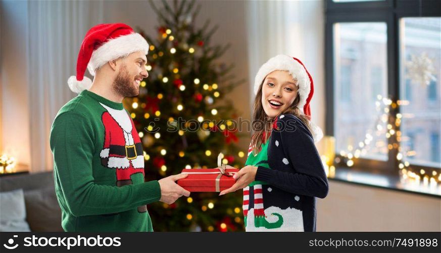 people and winter holidays concept - happy couple in santa hats with gift in ugly sweaters over christmas tree at home background. happy couple in ugly sweaters with christmas gift