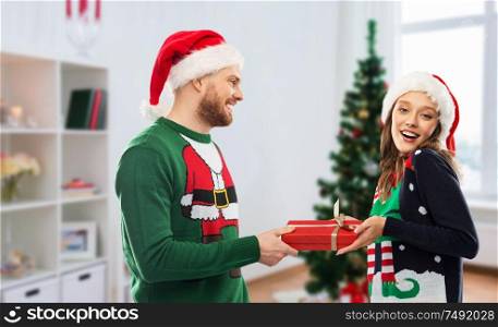 people and winter holidays concept - happy couple in santa hats with gift in ugly sweaters over christmas tree at home background. happy couple in ugly sweaters with christmas gift