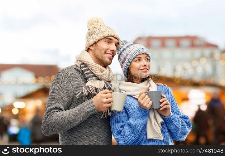 people and winter holidays concept - happy romantic couple in knitted hats and scarves with mugs over christmas market in old town of tallinn city background. happy couple drinking coffee at christmas market