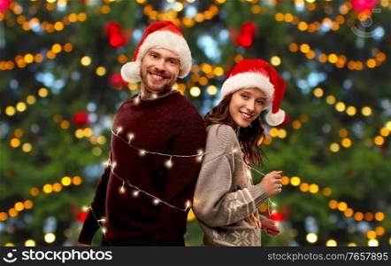people and winter holidays concept - portrait of happy couple in santa hats and sweaters tied with garland over christmas tree lights on background. happy couple in christmas sweaters and santa hats