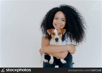 People, animal or pet care concept. Curly haired woman embraces favourite dog, smiles pleasantly, stands against white background, copy space area for your advertising content. Good friends.