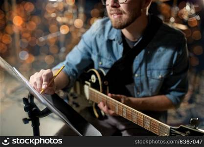 people, art and entertainment concept - man with guitar writing notes to music book at studio over holidays lights background. man with guitar writing to music book at studio