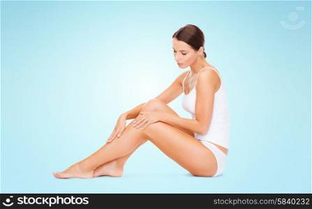 people, beauty and body care concept - beautiful woman in cotton underwear touching legs over blue background