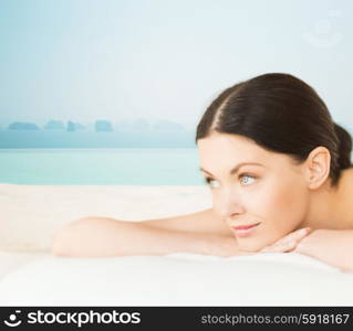 people, beauty and body care concept - happy beautiful woman lying on massage desk at spa resort over sea and infinity pool background