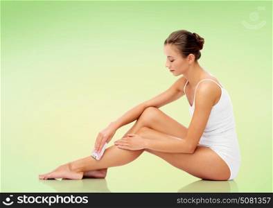 people, beauty and bodycare concept - beautiful woman with epilator removing hair from legs sitting on floor over green background. woman with epilator removing hair on legs
