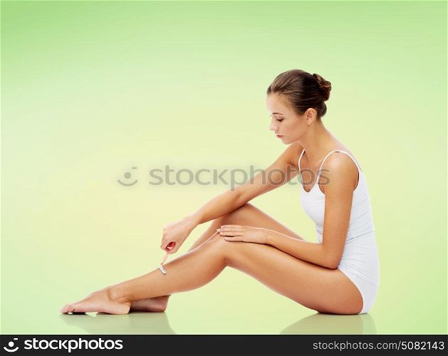 people, beauty and bodycare concept - beautiful woman with safety razor shaving legs sitting on floor over green background. woman with safety razor shaving legs