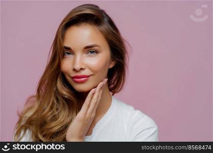 People, beauty and feminity concept. Lovely calm attractive lady with appealing look, keeps palm on cheek, satisfied after visit cosmetologist, has professional makeup, poses over purple background