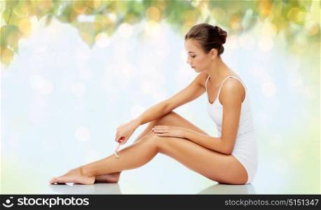 people, beauty and hair removal concept - beautiful woman with safety razor shaving legs sitting on floor over natural green background and lights. woman with safety razor shaving legs