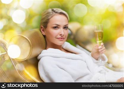 people, beauty and relaxation concept - beautiful young woman in white bath robe lying on chaise-longue and drinking champagne at spa over holidays lights background