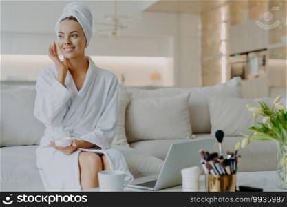 People beauty and skin care concept. Satisfied woman applies face cream enjoys facial treatments looks aside smiles gently uses cosmetic product poses at sofa in front of opened laptop computer