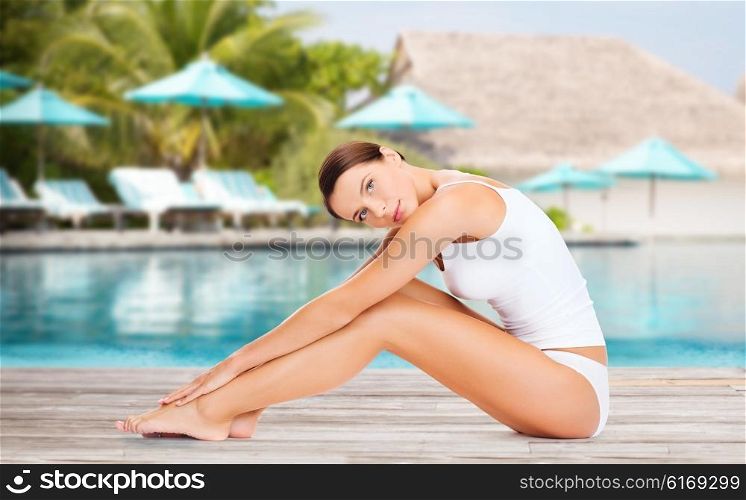 people, beauty and summer holidays concept - beautiful young woman in cotton underwear touching her legs over exotic hotel resort beach with swimming pool and sunbeds background