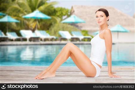 people, beauty and summer holidays concept - beautiful young woman in cotton underwear showing smooth skin of her legs over exotic hotel resort beach with swimming pool and sunbeds background