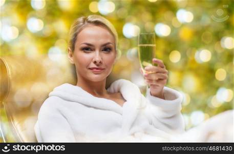 people, beauty, bodycare and relaxation concept - beautiful young woman in white bath robe lying on chaise-longue and drinking champagne at spa over holidays lights background