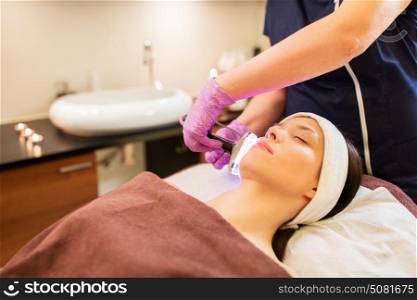 people, beauty, cosmetic treatment, cosmetology and technology concept - beautician with microdermabrasion device doing face exfoliation to young woman lying at spa parlor. young woman having face microdermabrasion at spa
