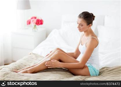 people, beauty, cosmetics, bodycare and spa concept - beautiful woman applying moisturizing cream to her legs at home bedroom. beautiful woman applying cream to her legs at home