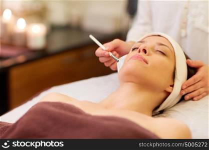 people, beauty, cosmetology and treatment concept - close up of beautiful young woman lying with closed eyes and cosmetologist applying facial mask by brush at spa. beautician applying facial mask to woman at spa