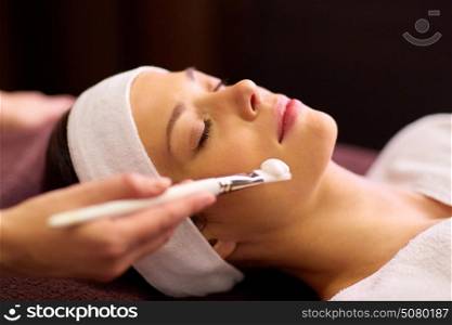 people, beauty, cosmetology and treatment concept - close up of beautiful young woman lying with closed eyes and cosmetologist applying facial mask by brush at spa. beautician applying facial mask to woman at spa