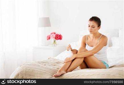people, beauty, depilation, epilation and bodycare concept - beautiful woman sitting on bed and with feather touching bare legs at home bedroom. woman with feather touching bare legs on bed