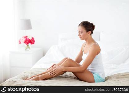 people, beauty, depilation, epilation and bodycare concept - beautiful woman touching smooth leg skin on bed at home bedroom. beautiful woman with bare legs on bed at home