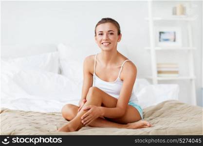 people, beauty, depilation, epilation and bodycare concept - beautiful woman touching smooth leg skin on bed at home bedroom. beautiful woman with bare legs on bed at home