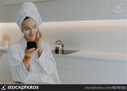People beauty hygiene concept. Adorable young woman in white bathrobe applies beauty patches under eyes for perfect healthy skin holds mirror looks thoughtfully aside poses over kitchen interior