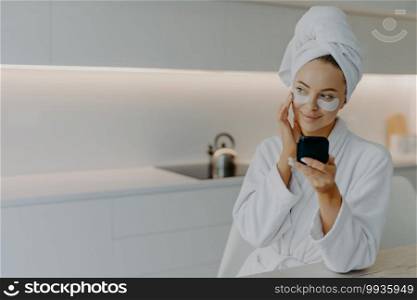 People beauty hygiene concept. Adorable young woman in white bathrobe applies beauty patches under eyes for perfect healthy skin holds mirror looks thoughtfully aside poses over kitchen interior
