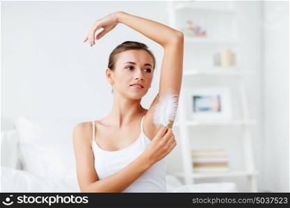 people, beauty, hygiene, depilation and epilation concept - beautiful woman sitting on bed and touching her armpit with feather at home bedroom. woman with feather touching her armpit at home