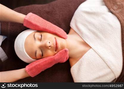people, beauty, lifestyle and relaxation concept - beautiful young woman lying with closed eyes and having face massage with terry gloves at spa. woman having face massage with terry gloves at spa