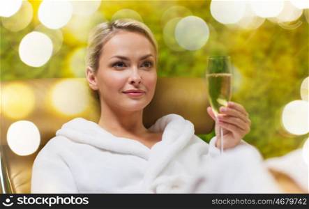 people, beauty, lifestyle, holidays and relaxation concept - beautiful young woman in white bath robe lying on chaise-longue and drinking champagne at spa over holidays lights background