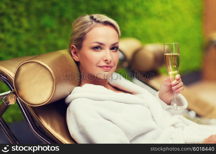 people, beauty, lifestyle, holidays and relaxation concept - beautiful young woman in white bath robe lying on chaise-longue and drinking champagne at spa