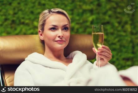 people, beauty, lifestyle, holidays and relaxation concept - beautiful young woman in white bath robe lying on chaise-longue and drinking champagne at spa