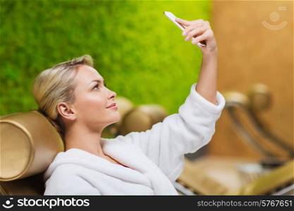 people, beauty, lifestyle, technology and relaxation concept - beautiful young woman in white bath robe taking selfie with smartphone at spa