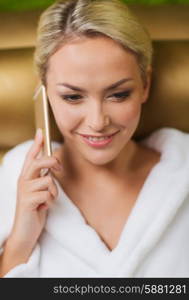 people, beauty, lifestyle, technology and relaxation concept - beautiful young woman in white bath robe with smartphone calling and talking at spa