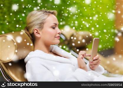 people, beauty, lifestyle, technology and relaxation concept - beautiful young woman in white bath robe with smartphone social networking at spa with snow effect
