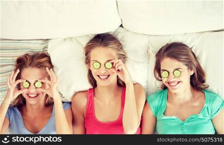 people, beauty, natural cosmetics and pajama party concept - happy friends or teenage girls with cucumber mask having fun and lying in bed at home. happy young women with cucumber mask lying in bed