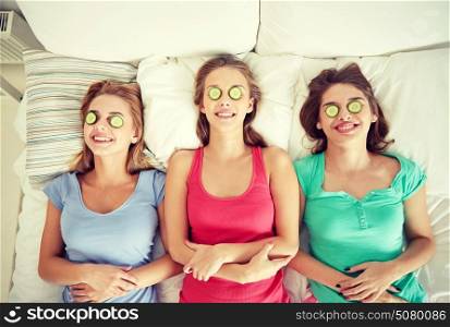 people, beauty, natural cosmetics and pajama party concept - happy friends or teenage girls with cucumber mask having fun and lying in bed at home. happy young women with cucumber mask lying in bed